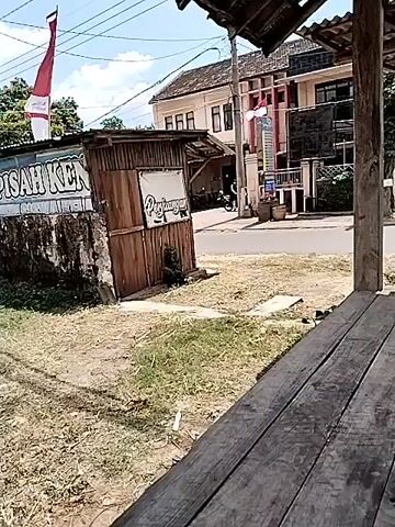 The video shows a street scene with a few buildings and a small wooden structure. In the foreground, there is a wooden platform or table. A person on a motorcycle rides past the wooden structure and down the street. Another person on a motorcycle follows shortly after. The environment appears to be an outdoor setting with grass and dirt on the ground. There are flags visible on one of the buildings.