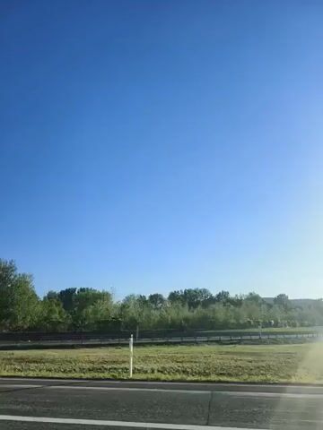 A wide shot captures a clear blue sky above a line of trees and vegetation. A road is visible in the foreground with sunlight glinting on the right side of the frame. The scene appears to be taken from a moving vehicle.