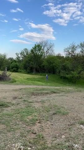 The video shows a person using a weed whacker to cut grass in a grassy area near trees. The person is wearing overalls and is seen from a distance. The camera then pans to the interior of a vehicle, showing the steering wheel and dashboard, with the view through the windshield showing the same outdoor scene. The sky is blue with white clouds.