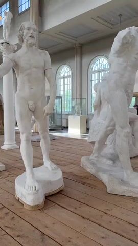 The video shows a room with many sculptures of nude men. The sculptures are all white and made of plaster. The room has high ceilings and large windows that let in natural light. The floor is made of wood planks. The sculptures are arranged in a line, and there are small signs in front of them.