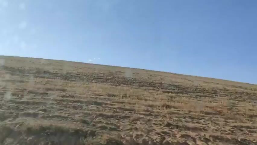The video shows a landscape view from a moving vehicle. The foreground is dominated by a dry, grassy slope with visible soil erosion and terracing. In the mid-ground and background, the slope continues upwards, covered in sparse, dry vegetation. The sky above is clear and blue with a few faint clouds. The camera appears to be positioned inside a vehicle, likely looking out a window, as indicated by a slight glare and streaks on the left side of the frame. The overall impression is of a dry, arid, or semi-arid environment under a bright sky.
