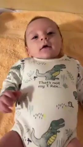 A baby lies on its back on a folded orange towel. The baby is wearing a white onesie that has a cartoon T. rex print and the words “That’s not my T. Rex.” The baby's head is centered in the shot, and its face is visible. The baby opens its mouth as if about to cry.