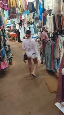 The video shows two women walking through a crowded outdoor market. The women are strolling through the market, past rows of clothes, bags, and various other items for sale. The market appears to be in an outdoor location with many stalls and a sandy ground.