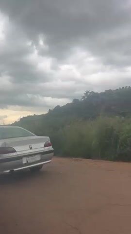 The video shows a side view of a silver sedan parked on a dirt road. The car is facing away from the camera and is partially obscured by vegetation. In the background, there is a hillside covered in trees and bushes. The sky is cloudy and overcast, suggesting it may rain soon. The camera pans to the right, revealing more of the hillside and a power line tower in the distance. A white van with advertising on its side is also visible.