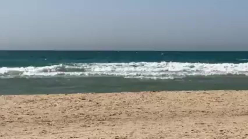 The video shows a sandy beach with three people swimming in the ocean. The water is turquoise with white waves. The sky is light gray. The sand is tan and undisturbed in the foreground.