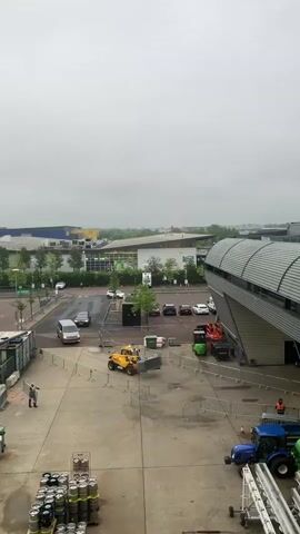 The video shows an outdoor industrial-type setting under a cloudy sky. In the foreground, there are stacks of kegs near the bottom left corner. A man wearing a safety vest walks across the paved area. A yellow front-end loader is moving a metal barrier. In the middle ground, a variety of vehicles are present including cars, vans, a forklift, and a blue tractor. In the background, there are various buildings and trees.