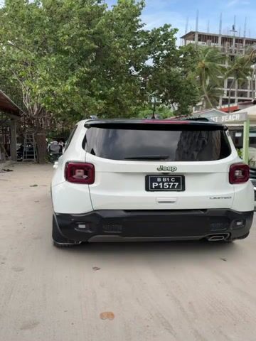 The video shows a white Jeep Renegade Limited parked on a sandy road. The car is facing away from the camera. The license plate reads "B B1 C P1577". A man walks into the frame from the right and towards the camera.  Behind the car are trees and buildings, including one with "Salt Beach" written on the side. There are also motorcycles parked along the side of the road. The sky is partly cloudy.