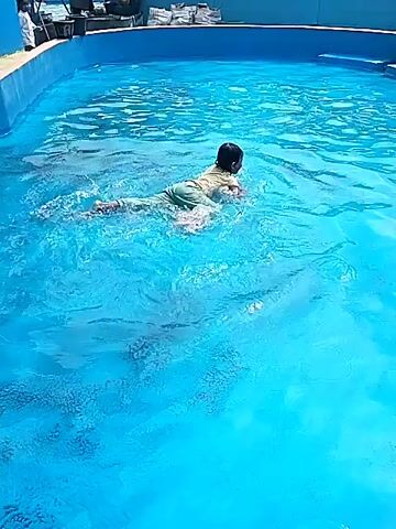 A child is swimming in a blue swimming pool. The child is wearing a light-colored shirt and green shorts. The child is facing away from the camera and appears to be swimming freestyle. The water is clear and blue, with some ripples on the surface. In the background, there are people sitting by the pool and some structures that appear to be part of a resort or recreational facility. The sky is visible and appears to be bright, suggesting it is daytime.