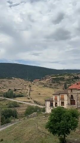A scenic view of a rural landscape under a cloudy sky. In the foreground, there is a house with an orange roof and a green tree. A road winds through fields and a small village can be seen in the distance. A large hill covered in trees forms the backdrop. There are no people visible in the video.