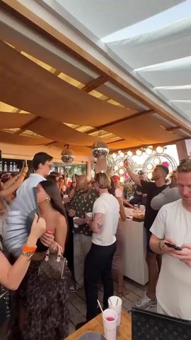 The video shows a crowd of people partying under a partially shaded outdoor structure. Most people are standing and moving around, some holding drinks. A few people are looking at their phones. The space is decorated with disco balls and colorful circular decorations in the background. Some people are wearing sunglasses. The atmosphere is lively and festive.