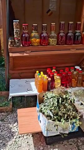 The video shows an outdoor market stall with various preserved foods and produce for sale. A wooden stall structure is visible, displaying rows of jars filled with fruits, vegetables, and possibly pickled items. These jars are arranged on shelves. Below the shelves, a table is set up with more jars of preserves, including what appears to be honey or jam, and bottles of liquids. In the foreground, several boxes are filled with fresh green apples. To the right, a woman sits on a chair behind a table, interacting with items on the table, including a mobile phone. Another person, partially visible, is seen working at the stall further down. The ground is covered with gravel. A Romanian flag is displayed on the wooden structure. The overall scene suggests a roadside market or farm stand.