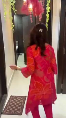 A woman is walking down a hallway. She is wearing a pink outfit with an orange floral print. The hallway is decorated with greenery and a pink light fixture. There is a brown doormat on the floor. The walls are beige and there are dark wooden doors on either side of the hallway. The woman dances slightly.