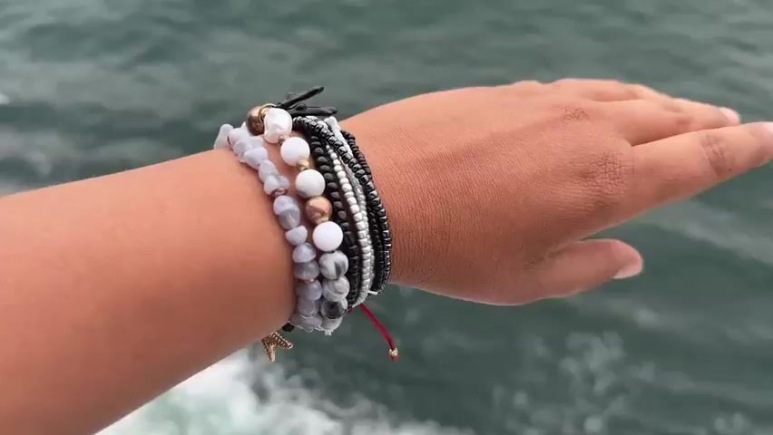 A hand adorned with multiple bracelets extends over a body of water. The bracelets are a mix of beaded strands with varying colors and materials, including white, gray, black, and metallic beads, some appearing to be stacked together. The hand then moves to extend its palm over the water. The water is a dark, greenish gray color, with subtle ripples and waves visible on the surface. The background is predominantly the ocean, filling the frame and creating a sense of openness.