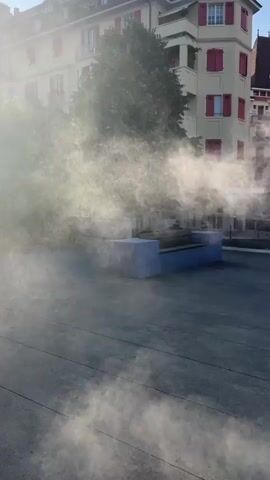 The video shows an outdoor scene with a building in the background and a street or paved area in the foreground. A significant amount of mist or fog is present, obscuring some parts of the scene. In the middle ground, there appears to be a low structure, possibly a bench or seating area, that is partially covered by the mist. Trees are visible behind this structure. The building in the background is multi-story with red-tiled roofs and balconies, and appears to be residential or commercial. The overall impression is an urban environment with an atmospheric condition creating a hazy or misty effect.
