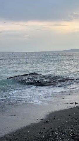 The video shows a view of the ocean from a beach.  The sky is overcast.  Waves are rolling in towards the beach and washing over a partially submerged wooden structure that appears to be a raft.  The beach is made up of gray and black sand and small rocks.  In the distance, a land mass can be seen along the horizon.