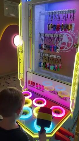 A young boy stands in front of a brightly lit arcade game called "Whack-a-Mole." The game features a display of small keychain toys hanging on the right side, and a panel of colorful, illuminated holes at the bottom. The boy is holding a red, yellow, and blue toy hammer and is actively playing the game, swinging the hammer at the holes where characters pop up. Another person, partially visible to the left, is also watching or possibly waiting to play. The environment appears to be an indoor entertainment space, likely an arcade or amusement center, with soft lighting and patterned flooring.