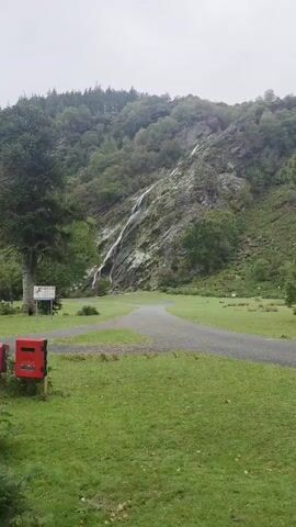 The video shows a view of a waterfall cascading down a rocky hillside. The surrounding landscape is lush and green, with trees and grass covering the lower slopes. There is a paved path winding through the grassy area in the foreground, and what appears to be a picnic table is visible in the distance. The sky is overcast, suggesting a cloudy day. The camera pans slowly from left to right, capturing different angles of the waterfall and its surroundings. There are no people visible in the video.