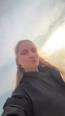 A young woman with blonde hair and wearing a black jacket records a selfie video on a beach. In the background, the sea laps at the shore, and two figures are visible. One is bent over near the water’s edge, while the other, appearing to be a small child in an orange jacket, walks along the beach. The sun is shining brightly, creating a hazy effect on the horizon. The woman’s face is the main focus of the shot, and she looks directly at the camera with a neutral expression.