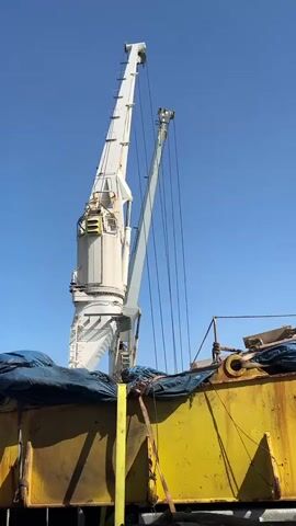 A time-lapse video shows a large white crane against a clear blue sky. The crane's boom arm is angled upwards and extends diagonally across the frame. Below the crane, a large, weathered yellow object, possibly a container or cargo, is partially visible, covered with blue tarps and straps. The crane appears to be in operation, with its cables taut and extended. The video captures the slow, steady movement of the crane.