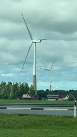 The video shows a landscape scene with two large wind turbines in the foreground and middle ground, respectively. The wind turbines are white with cream-colored bases, and their blades are in motion. In the background, there are several houses with red roofs nestled among trees. A road with a concrete barrier separates the foreground grassy area from the houses and wind turbines. The sky is overcast with dramatic, gray clouds. The overall mood is serene and somewhat dramatic due to the cloudy sky. The time of day appears to be daytime, likely afternoon or late morning, given the ambient light. The location is Koogu, Estonia, which suggests a rural or semi-rural setting typical for wind farm installations. There is no visible text in the video. The primary subjects are the wind turbines, the rural landscape, and the houses. The activity taking place is the rotation of the wind turbine blades, indicating that they are generating electricity.