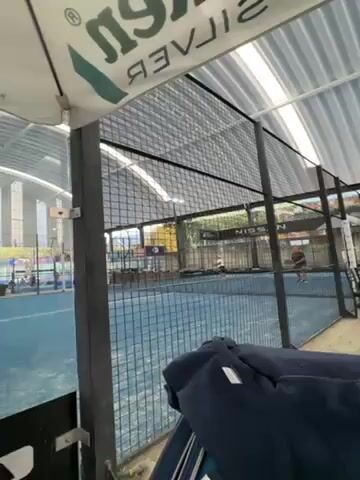 Here is a description of the video:

The video shows two people playing a game of paddle tennis on a blue court surrounded by black wire mesh. The court is indoors under a white metal roof, with one side open to the outdoors. There are ads visible in the background, including a Nissan sign. The video is taken from behind the court, with a blue jacket in the foreground partially obscuring the view. The camera angle is low and looking upwards, capturing the players' movements as they hit the ball back and forth.