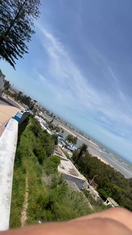 The video shows an elevated view of a coastal landscape on a bright, sunny day. The sky is blue with scattered, wispy clouds. Below, a sandy beach meets the ocean, with gentle waves lapping the shore. The ocean extends to the horizon. A line of buildings and structures runs along the edge of the beach in the distance.

In the foreground, lush green trees and vegetation cover the slope leading down towards the beach. Several buildings are visible among the trees, including some with flat roofs and others with more traditional architectural features. One building appears to have a satellite dish on its roof. The camera pans slowly, revealing more of the coastline and the buildings nestled among the greenery.