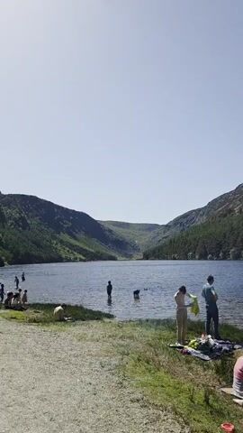 The scene depicts a lake surrounded by hills with lush green forests. Several people are engaging in different activities near the water. Some are wading into the lake, while others are sitting or standing on the grassy or gravelly bank. There is a dog swimming in the lake. The weather appears sunny, with a clear blue sky. The scene conveys a sense of relaxation and enjoyment of the outdoors.