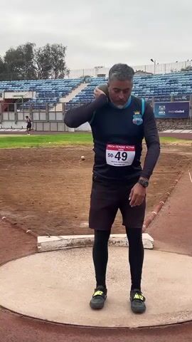A middle-aged man with graying hair and a beard, wearing a dark blue long-sleeved shirt with a light blue stripe, black shorts, black leggings, and black athletic shoes, stands in a shot put circle. He is holding a shot put against his neck with his right arm, preparing to throw. He has a race bib numbered 49 on his shirt. The background shows a dirt field, stadium seating with blue chairs, and trees under an overcast sky. Another person, a runner, is visible in the distance. The man appears focused on his throw.