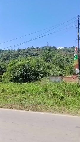 The video shows a view from a moving vehicle of a rural landscape under a clear blue sky. The road in the foreground is paved and appears to be a single lane in each direction. To the left of the road, there is a small, modestly sized house with a brown roof and light-colored walls. The house is situated on a slightly elevated piece of land covered with green grass and some scattered dry vegetation. Behind the house and to the right, the terrain rises, covered in dense green trees and foliage, forming a hillside. Power lines are visible stretching across the upper part of the frame, supported by utility poles. The overall scene suggests a tranquil, natural environment in what appears to be a developing or rural area.