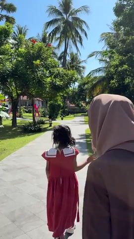 A young girl wearing a red dress and a woman in a hijab are walking on a paved path surrounded by lush greenery and palm trees. The girl is in the foreground, facing away from the camera, and appears to be looking ahead. The woman is behind her, also facing away. The sun is shining brightly, casting shadows on the path, suggesting it is a warm day. The environment appears to be a resort or a park-like setting.