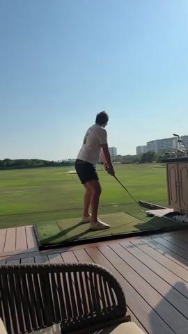 A man in a white t-shirt and dark shorts is swinging a golf club at a driving range. He is standing on a wooden deck with a green mat in front of him, and several golf balls are visible next to the mat. Behind him is a well-manicured green field, with buildings and trees visible in the distance under a clear, sunny sky. The man takes a full swing with the club and then holds his follow-through pose. The camera then switches to a close-up of the man's face, as he looks into the camera with a slight grimace. He is wearing a green baseball cap.