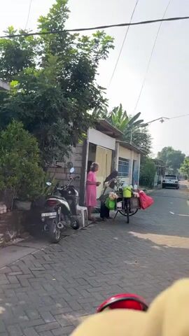 The video shows a street scene with two women and a bicycle cart. One woman, wearing a pink dress, stands on the left side of the frame, facing a building. The other woman, wearing a headscarf and a light-colored top, is behind a bicycle cart, interacting with the first woman. The cart is laden with various items, including a green bucket, plastic bags filled with produce, and what appears to be snacks or food items. The building behind them has a simple facade with a door and windows. To the left of the building, a motorcycle is parked. The street is paved with bricks, and trees and other vegetation are visible in the background. The sky is bright, suggesting daytime.