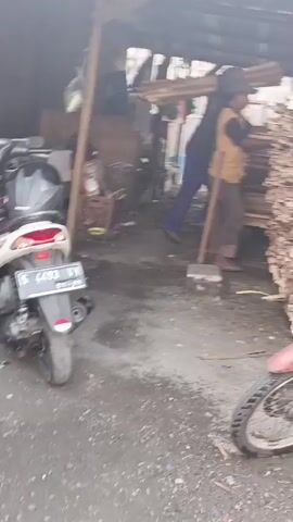 The video shows a person standing in what appears to be a lumberyard or wood workshop. The person is wearing a yellow vest and a hat, and is interacting with a large stack of lumber. There is a motorcycle parked nearby. The environment is somewhat cluttered and appears to be an outdoor or semi-outdoor setting with a roof structure overhead. The lighting is dim.