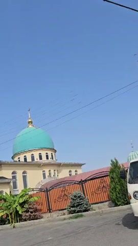 The video shows a mosque with a light yellow facade and a bright turquoise dome topped with a golden finial. To the left of the mosque is a tall, slender minaret, also light yellow, with decorative banding and a small pointed top. A fence with ornate black metal railings and a reddish-brown top encloses the mosque grounds. Several evergreen bushes and a banana tree with large green leaves are visible in front of the fence. The sky is clear and blue with a few faint horizontal lines, likely power lines, stretching across it. The mosque and minaret appear to be leaning slightly to the right. The footage is recorded from a low angle, looking upwards.