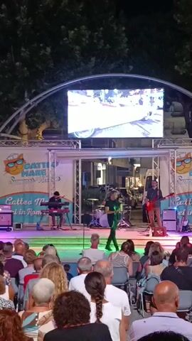 A live band performs on a stage outdoors at night, with an audience seated in front of them. The stage is set up with musical instruments including drums, keyboards, and guitars. A large LED screen is mounted above the stage, displaying images. The band consists of several members, including a vocalist, a guitarist, a keyboardist, and a drummer. The audience members are of various ages and appear to be enjoying the performance. The background shows trees and buildings, suggesting an urban or town setting. The atmosphere is festive, with colorful stage lighting illuminating the scene.