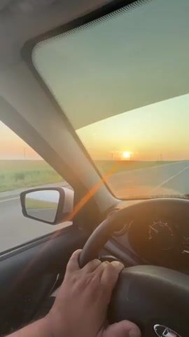 The media captures a first-person perspective from inside a car, with the driver's hands on the steering wheel. The view is directed forward through the windshield, showing a road stretching towards the horizon. The time of day is clearly sunset, with the sun a bright orb low in the sky, casting a warm, golden glow. The sky transitions from a pale yellow near the horizon to a soft green at the top of the frame, with a lens flare streaking across the image.

The setting appears to be a rural or open landscape, possibly agricultural land, suggested by the flat terrain and fields visible to the left of the road. In the distance, silhouetted against the bright sky, are several wind turbines, indicating a location with wind energy infrastructure. The road itself is paved and appears to be a highway or a major road, with a lane line visible. The car's dashboard is partially visible on the right, showing a speedometer and what appears to be a digital display with numbers "21:04" and "80", possibly indicating the time and speed.

The overall mood is serene and peaceful, evoking a sense of journey and the beauty of nature at dusk. The sunlight bathing the scene creates a tranquil atmosphere, suggesting a calm drive through the countryside as the day ends. The location context provided, Panika, Russia, helps to place this scene in a specific geographical area, though the visual cues themselves are general to many open, rural landscapes with wind farms.