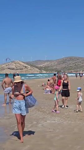 This is a vertical video of a crowded beach on a sunny day. The sky is clear and blue, and there are hills in the background with wind turbines. The beach itself is sandy and wet, with shallow water lapping at the shore. Several people are visible, most of whom are wearing swimwear. In the foreground, a woman with a straw hat and denim shorts is looking at her phone. Behind her, there are other beachgoers, including men and women, some of whom are standing in the water. Further in the background, there are more people, some sitting on the sand and others wading in the water. There are also several kites in the air, suggesting that kitesurfing or kiteboarding is taking place. Some of the kites are large and colorful, while others are smaller. The overall impression is one of a lively and active beach scene on a summer day.
