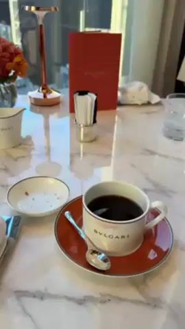 The video shows a table setting with a cup of coffee, a small plate with crumbs, and a creamer jug labeled "Dolci". A Bulgari menu stands behind the setting. The camera then pans to a window revealing a view of a building with a grid-like facade. The shot widens to reveal a high-ceilinged room with large windows, tables with floral arrangements, and a Christmas tree in the background. The room appears to be a restaurant or cafe with modern decor.