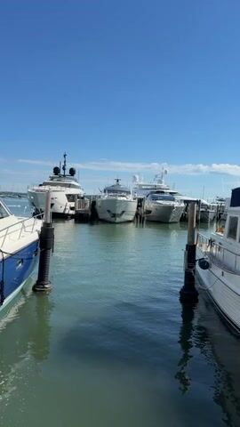 The video shows a marina on a clear, sunny day. Several yachts and boats are docked. In the foreground, a white boat with a blue stripe is docked on the left, and another white boat is docked on the right. In the background, three large white yachts are docked side-by-side. The water is calm and reflects the sky and the boats. The sky is a clear blue with a few wispy clouds. There are wooden docks and mooring posts visible.