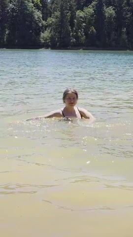 A young woman in a black swimsuit is swimming in a lake. She is in the center of the frame and is splashing the water with her hands. The water is a murky yellow-green color. Behind her, the lake stretches out to a shoreline lined with green trees. The sky is visible above the trees. The video is shot from a low angle, looking up at the woman.