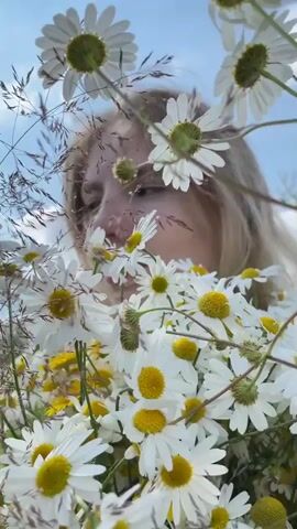 A person with blonde hair is holding a bouquet of daisies in front of their face, obscuring most of their features. The daisies are white with yellow centers and are the main focus of the foreground. The background is out of focus, showing a blue sky with some clouds. The person's eyes are visible and looking slightly downwards. The lighting suggests it is daytime, possibly outdoors.