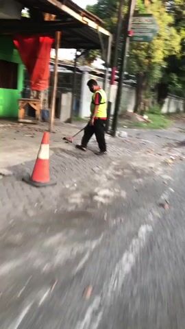 The media file shows a person sweeping the street. The person is wearing a yellow safety vest and black pants. They are using a broom and appear to be working on a paved area. In the foreground, there is an orange traffic cone. Behind the person, there is a building with a green wall and a red cloth hanging from an overhang. Trees and other vegetation are visible in the background, suggesting an outdoor setting. The lighting suggests it might be daytime, possibly early morning or late afternoon, given the shadows and the quality of light. The scene appears to be a street or pathway in a town or village. There is some debris on the ground. The motion blur in the image indicates movement. The location context provided is Ngampelsari, Indonesia. No visible text is discernible in the media file.