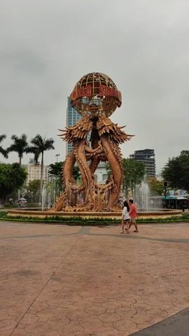 The video shows a large fountain in a square. The fountain features two large, golden dragon sculptures entwined and supporting a metal globe with the words "WORLD SUN" on it. Water sprays from multiple points around the base of the fountain. A couple, a woman in white and a man in red, walk past the fountain. The square is paved with reddish-brown bricks. In the background, there are buildings, palm trees, and a cloudy sky. Other people can be seen in the periphery of the shot.