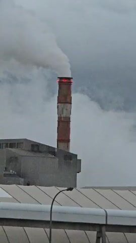 The video shows a tall, cylindrical smokestack emitting a large plume of white smoke against a gray, overcast sky. The smokestack has alternating red and white bands, with a red glowing band at the very top. To the left of the smokestack, a portion of a gray industrial building is visible. In the foreground, a section of a white, flat roof or structure with a curved edge is present, and a black lamp post with a curved arm extends upwards. The overall impression is of an industrial scene with active emissions.