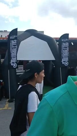 The video shows an outdoor event, possibly a motorcycle convention or fair, on a partially cloudy day. The scene begins with a woman in white pants, a black top, and a red cap standing in front of an inflatable black archway. A flag to her side displays the word "Motocicletas." The camera then pans to reveal a man in a grey shirt, a person in a black jacket and cap, and several motorcycles parked nearby. Subsequently, a group of individuals is seen walking amidst the motorcycles, including a young man in a beige shirt. The scene concludes with two individuals shaking hands, positioned among a crowd and the parked motorcycles. The environment includes a building with a tiled roof and palm trees in the background. Loud music is playing during the event.