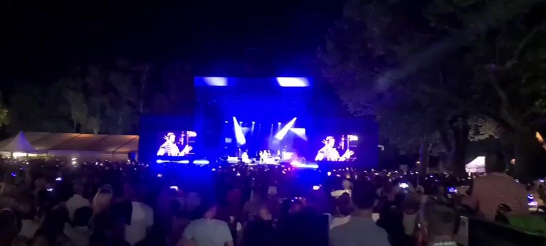 The video captures an outdoor night concert with a large crowd gathered in front of a brightly lit stage. The stage features multiple spotlights and large screens displaying close-ups of performers. The audience, a dense mass of people, is illuminated by the stage lights and many are holding up their phones, presumably to record the performance. The background is dark, suggesting the concert is taking place in a park or open area with trees visible on the right side. The atmosphere appears lively and engaging.