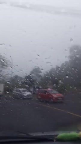 The video, shot from inside a car, shows a road on a rainy day. Raindrops are visible on the windshield. The road is wet and there are cars and trucks driving on it. Trees line the side of the road, their branches bare. The sky is overcast and gray, giving the scene a gloomy feel. In the foreground, parts of the car's dashboard are visible. The audio includes music, suggesting someone in the car is playing it.