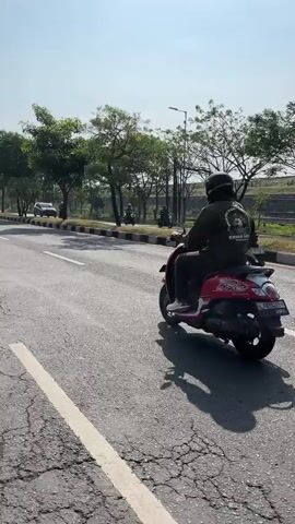 A person wearing a helmet and a dark green jacket with a circular graphic on the back is riding a red scooter on an asphalt road. The road has white lane markings and appears to be cracked in places. The rider is facing away from the camera. To the left of the road, there is a sidewalk or median strip with trees and a few other motorcycles or scooters parked or being ridden in the distance. A white car is also visible on the road in the far distance. The sky is clear and bright, suggesting it is daytime with sunny weather. The scene appears to be outdoors in a semi-urban or suburban environment.