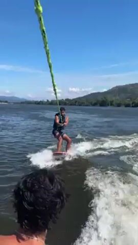 The video shows a man wake surfing behind a boat on a lake on a sunny day. The wake surfer is wearing a life vest and holding onto a rope. He is riding the wake created by the boat. The person filming is on the boat, and at one point, the camera pans to show the interior of the boat, including the dashboard and a wakeboard. In the distance, there are mountains and trees. Another boat can be seen briefly on the lake.