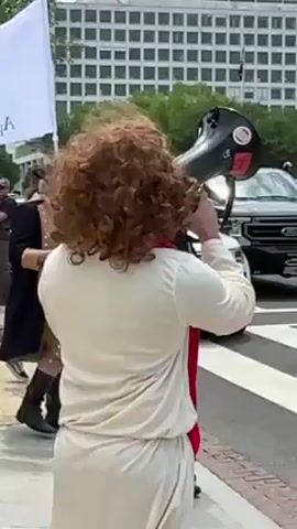 The video depicts an outdoor scene on a sunny day in Washington, United States.

Initially, a person with curly reddish-brown hair, wearing a light-colored garment and a red sash, is seen from behind. They are holding a black megaphone to their mouth, actively speaking. Behind them, a white flag with green text, where "Appeal" is partially visible, is displayed. A dark-colored Ford pickup truck drives past in the street, with a large, multi-story building visible in the background.

The camera then shifts focus to a street with a prominent crosswalk. A person wearing a red hat and a dark shirt walks across the zebra crossing. They briefly extend their arms outwards before continuing to walk. A white extended van or limousine is parked on the side of the road. Green trees line the street, and the same large building is visible further down. The presence of a megaphone and flag suggests a public gathering or protest event. The weather appears clear and bright.